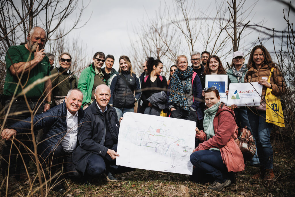 Gruppenfoto, in der Mitte wird Plan gehalten, und Schilder von anderen Personen, inmitten einer Hecke
