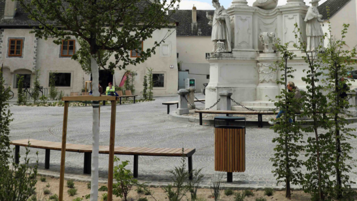 Platz mit Staudenbeet, historischem Monument im Hintergrund, im Vordergrund Staudenbeet mit Baum und Sträuchern