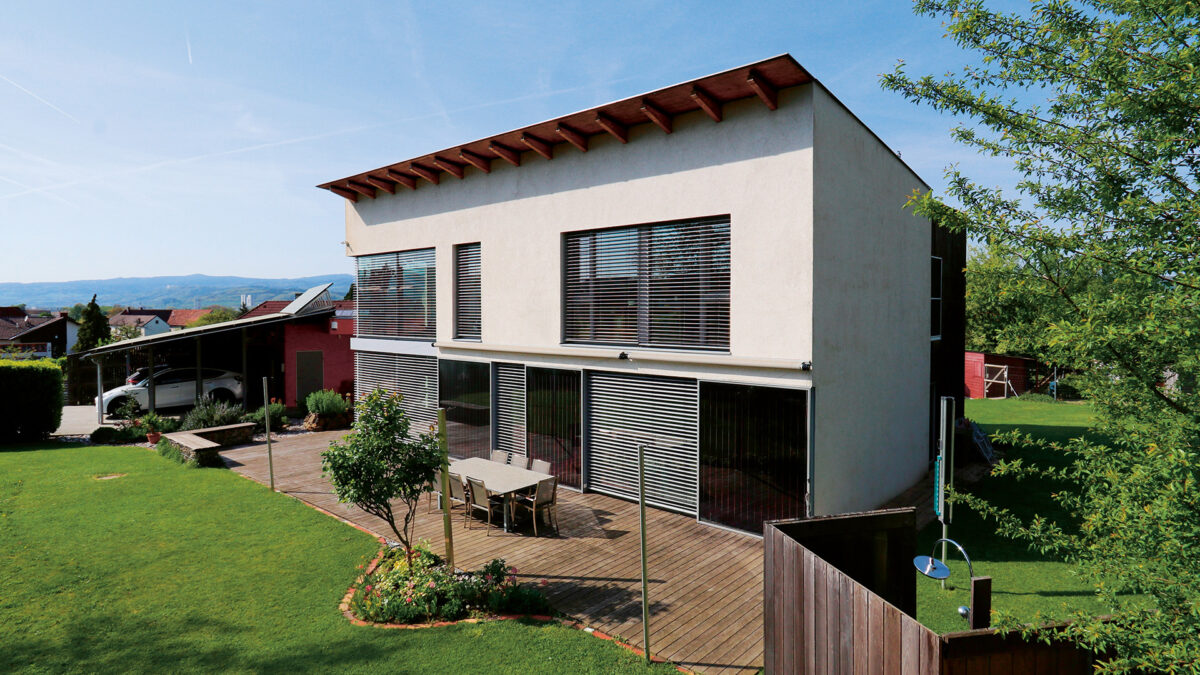 Helles Haus mit grüner Wiese rundherum, Terrasse mit Holz, kleine Bäume.