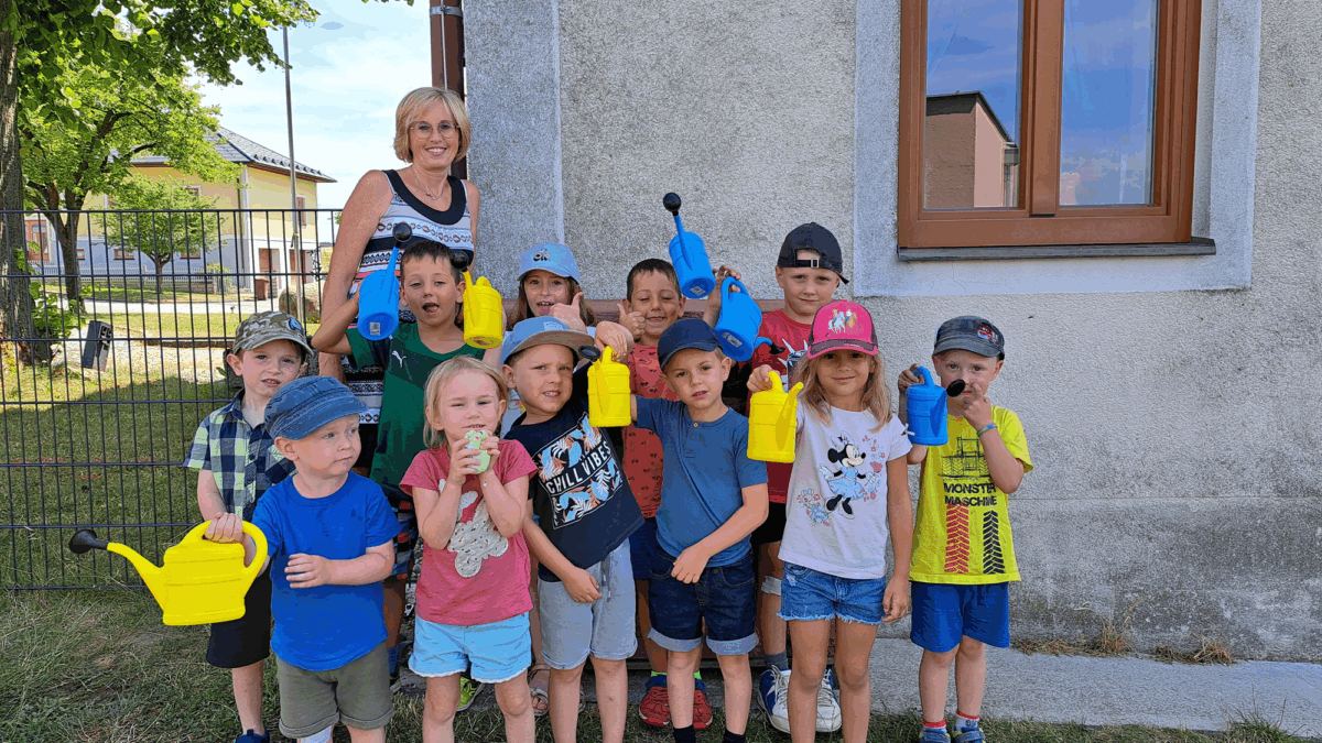 Eine Gruppe Kinder mit Gießkannen in der Hand vor einem Haus mit Pädagogin