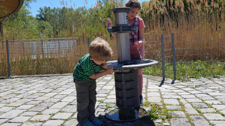 Zwei Kinder bei Trinkbrunnen, eines trinkt.