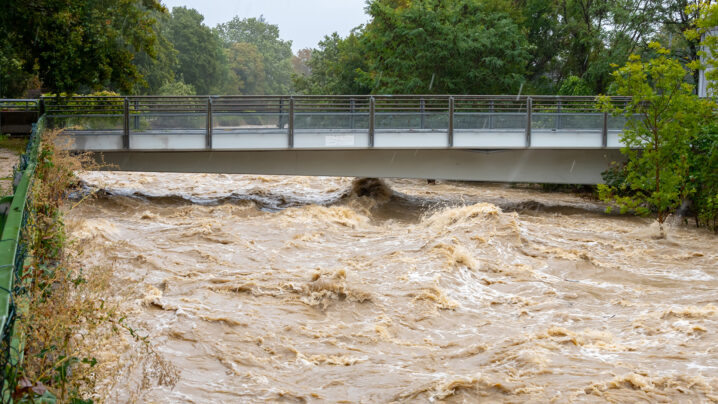 Brücke, braunes aufgewühltes Flussgewässer, es regnet, grüne Vegetation am Ufer