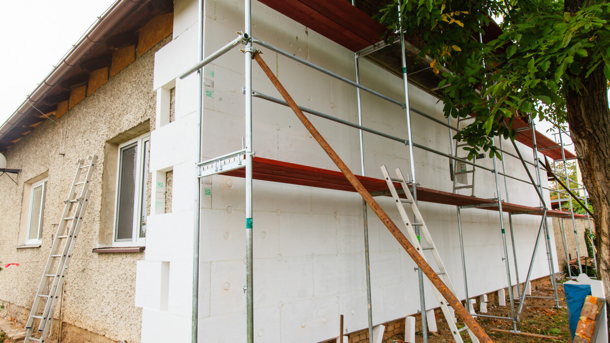 Altes Haus, auf einer Seite eingerüstet und Dämmmaterial angebracht, Gerüst und lehnende Leitern an der Fassade, daneben Baum