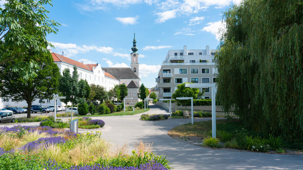 vorne links Staudenbeete, rechts eine grüne Weide, Wege mit moderner Straßenbeleuchtung, im Hintergrund ein Kirchturm und ein modernes Wohnhaus.
